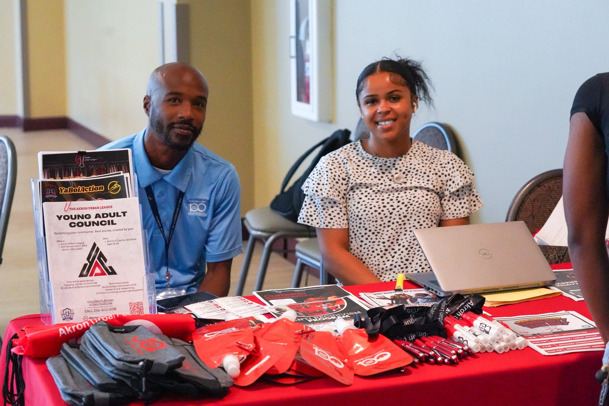 Two young adults smiling behind a table with flyers, pens, and red swag, including one titled "YOUNG ADULT COUNCIL."